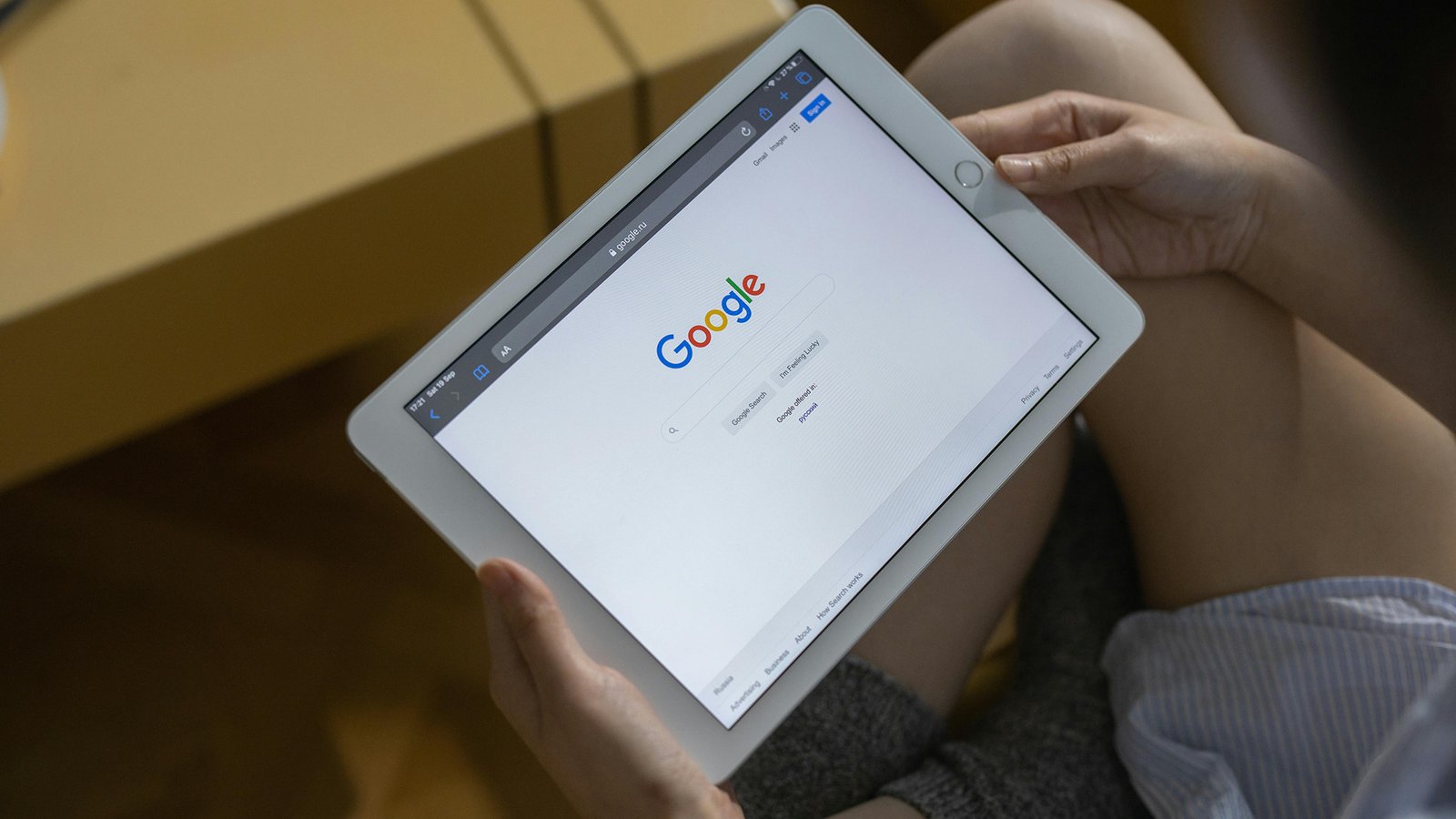 Individual holding a white tablet displaying Google homepage with search bar, seated near a wooden surface