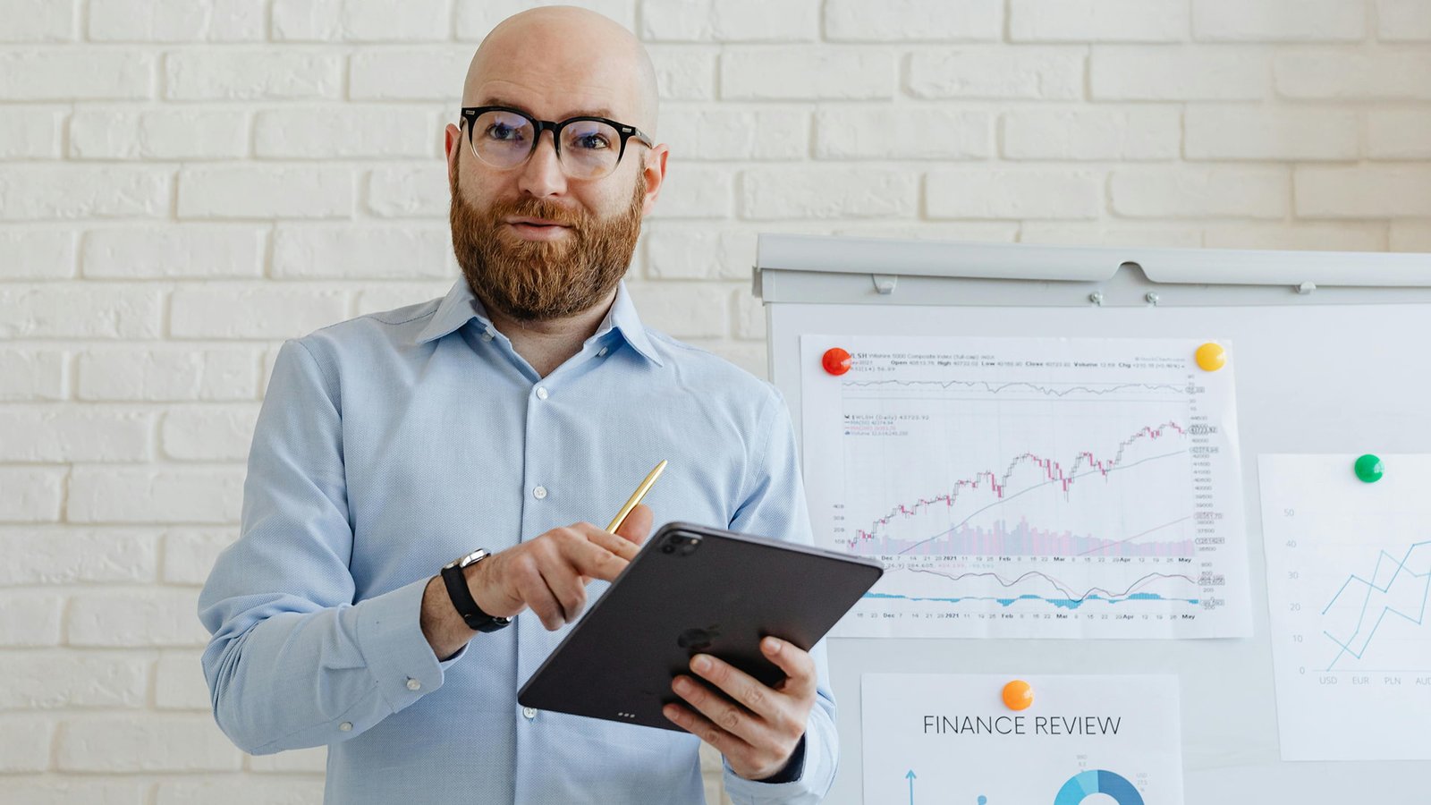 SEO expert holding a tablet while standing near a financial review board with charts and analytics in an office setting.